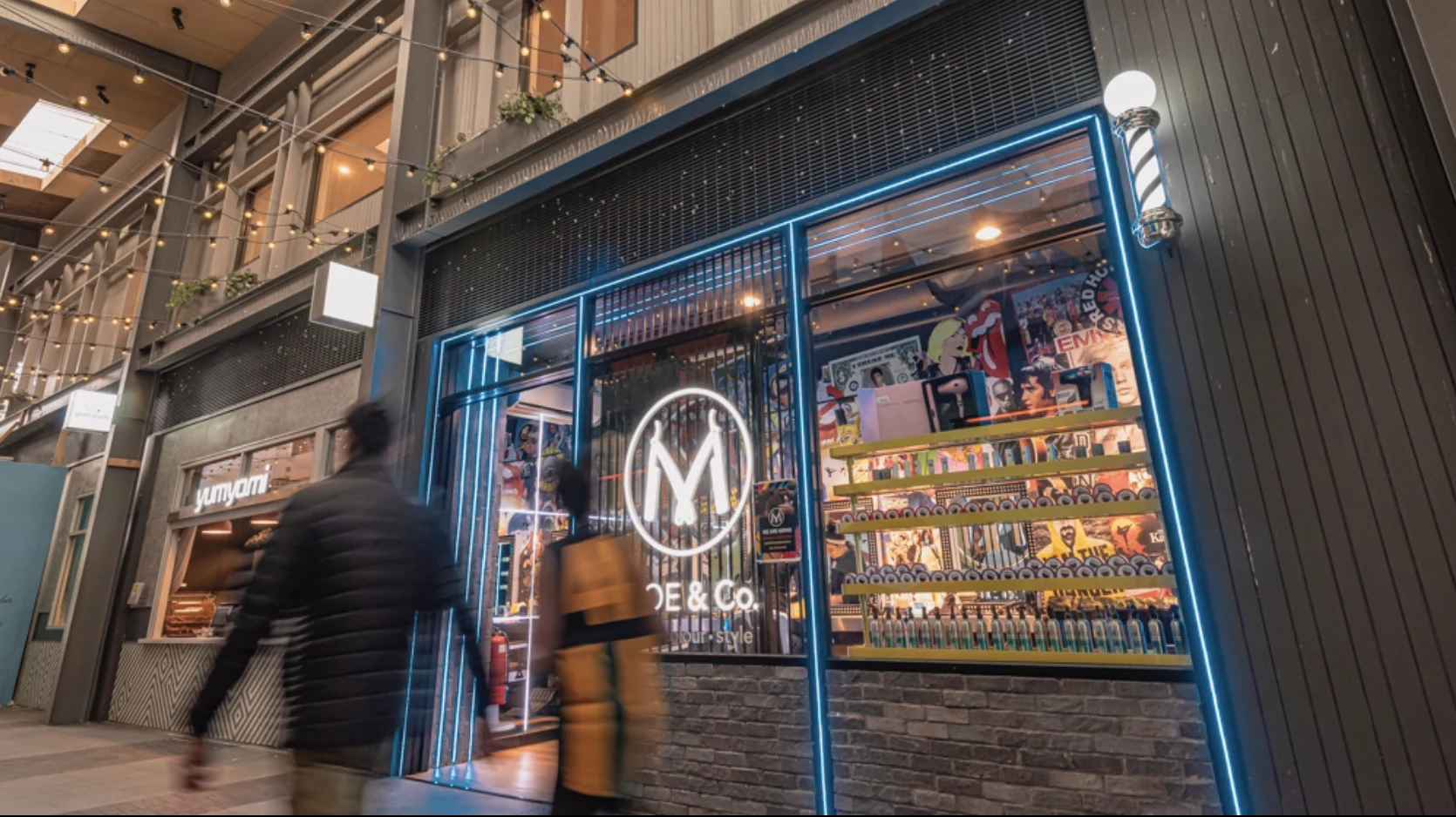 MOE & Co. Camberwell Place shopfront — the covered Burke Road laneway, festoon lights, blue neon, chrome barber pole and the poster wall glowing through the glass