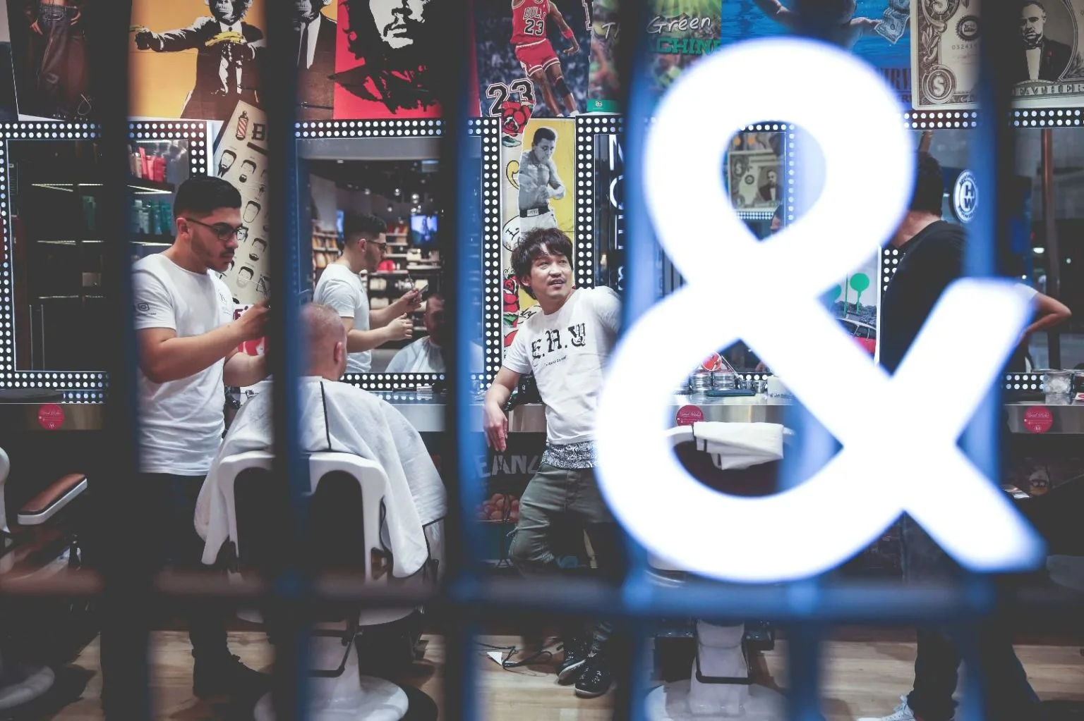 Inside MOE & Co. — shot through the shopfront: the lit ampersand sign, Jordan and Ali on the wall, a stylist mid-cut and mid-laugh