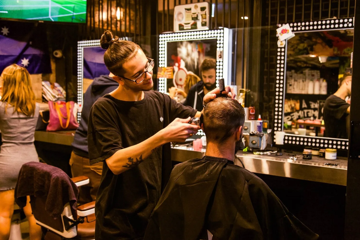 Clipper fade in progress at a bulb-framed station — a stylist working with a client