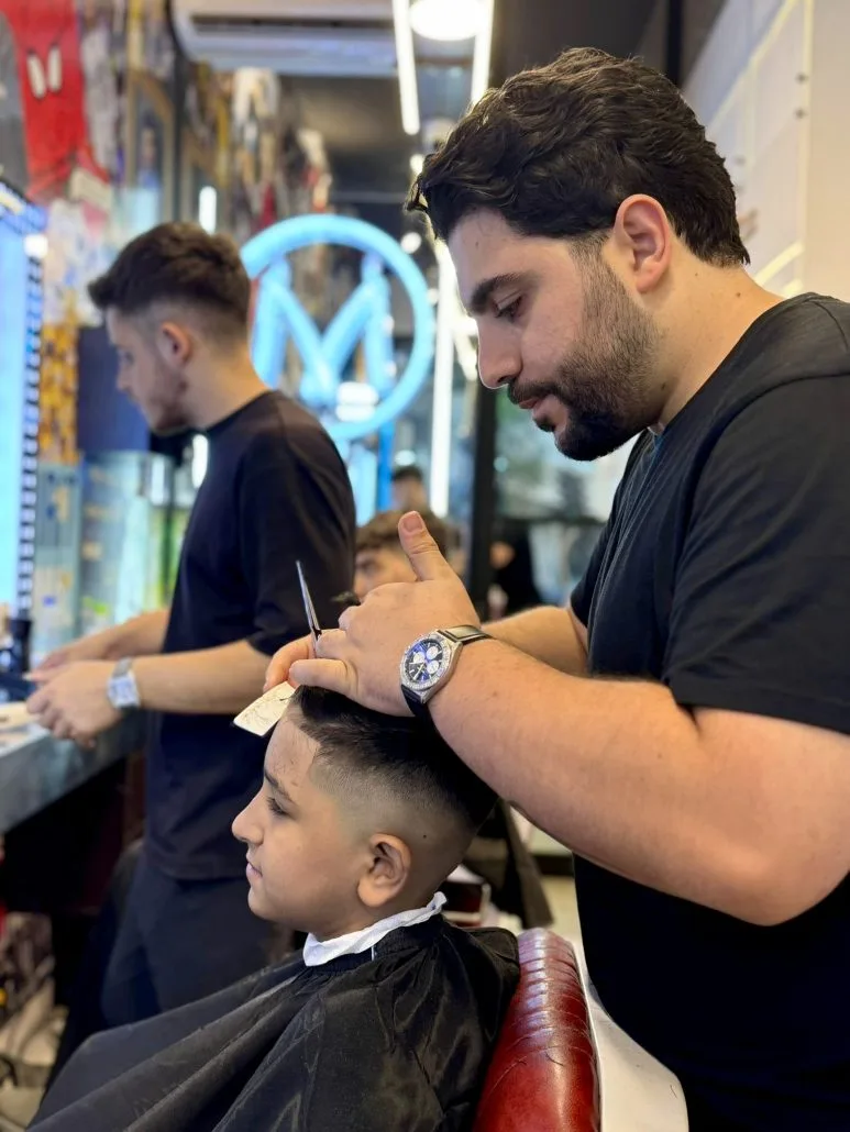 A young client in the chair — a stylist at work beneath the blue neon M, both focused