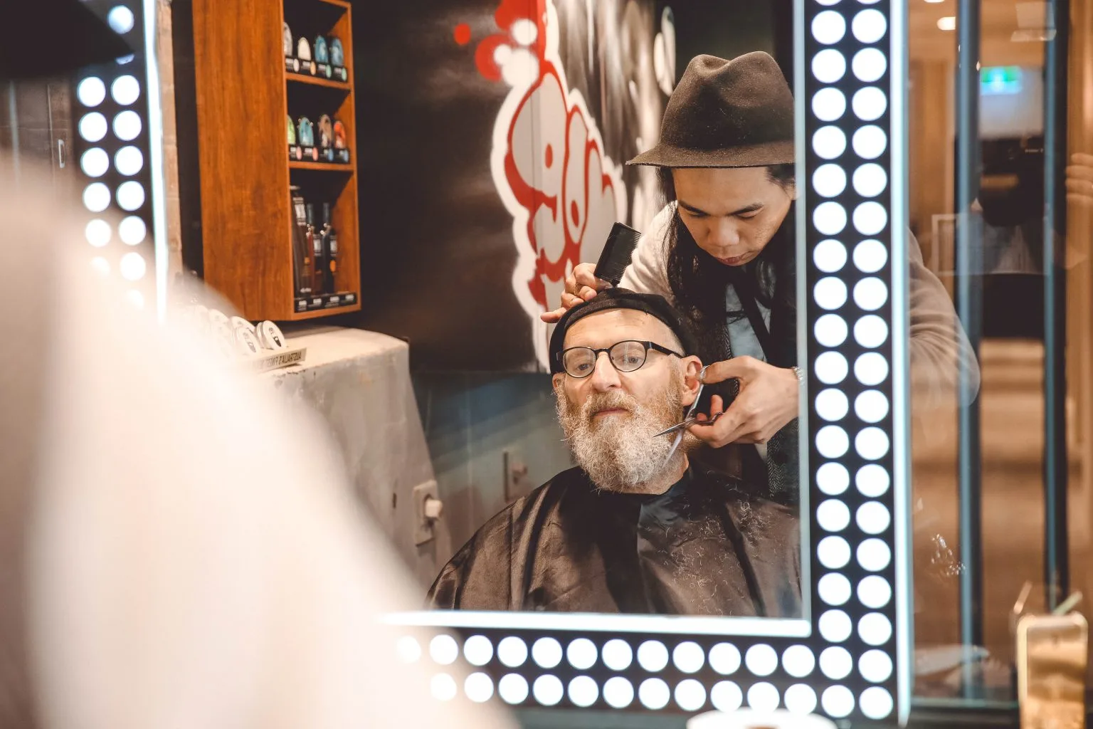 A stylist scissor-cutting a client at a bulb-framed mirror — calm, precise, unhurried